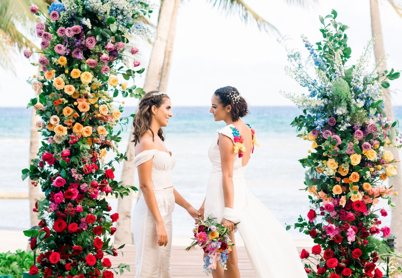 Two women in wedding attire stand holding hands under a colorful floral arch by the ocean, with palm trees in the background.