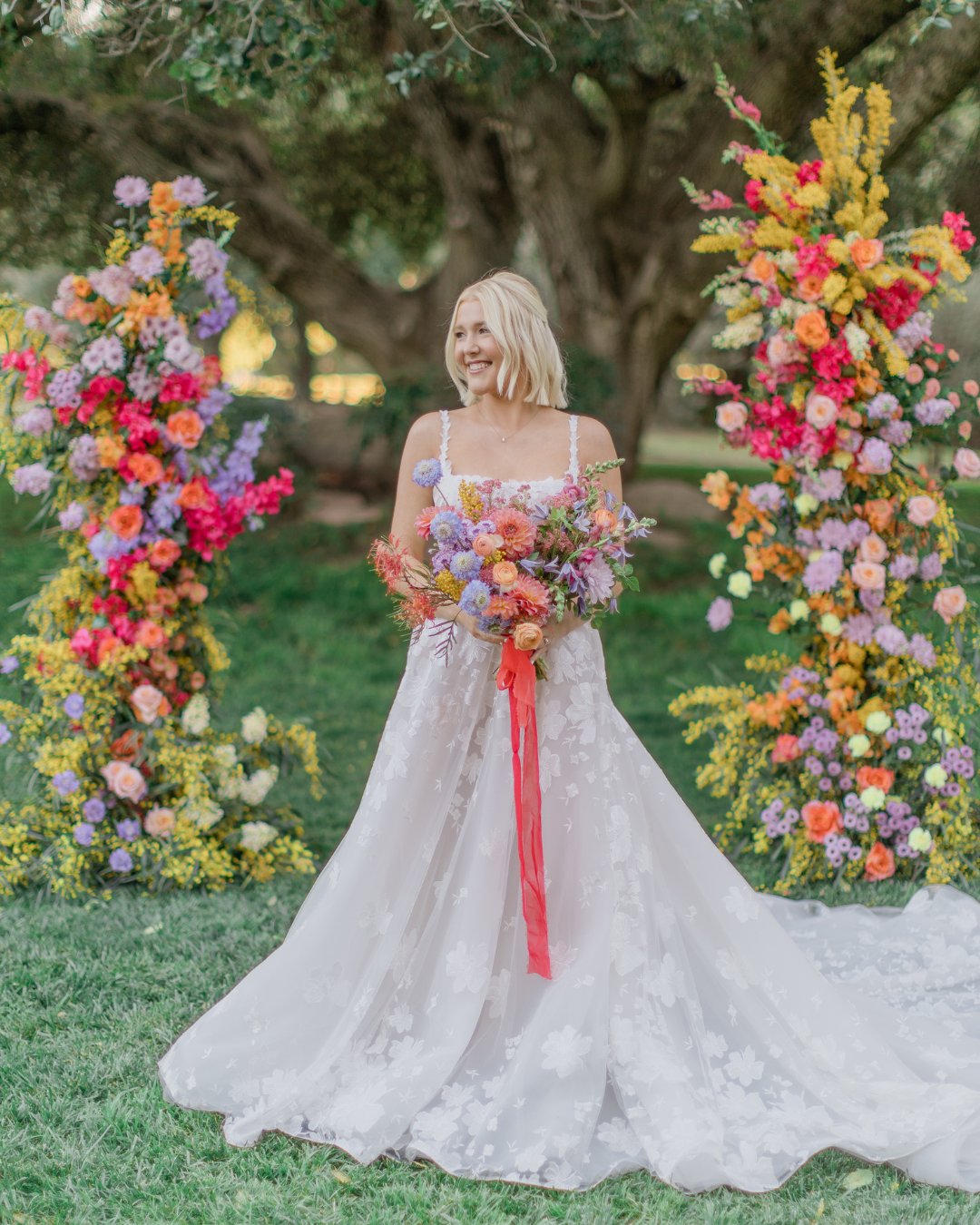 A bride in a white floral gown holds a colorful bouquet, standing between two vibrant floral arrangements in an outdoor setting.