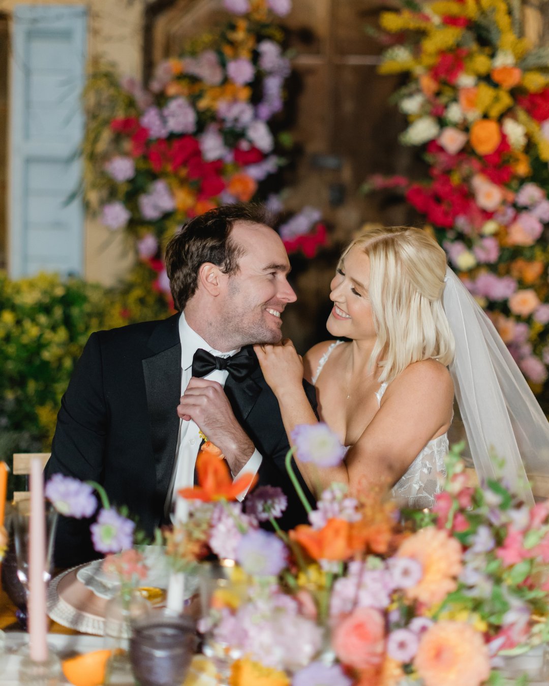 A bride and groom in formal attire sit closely at a decorated table with colorful flowers, smiling at each other.