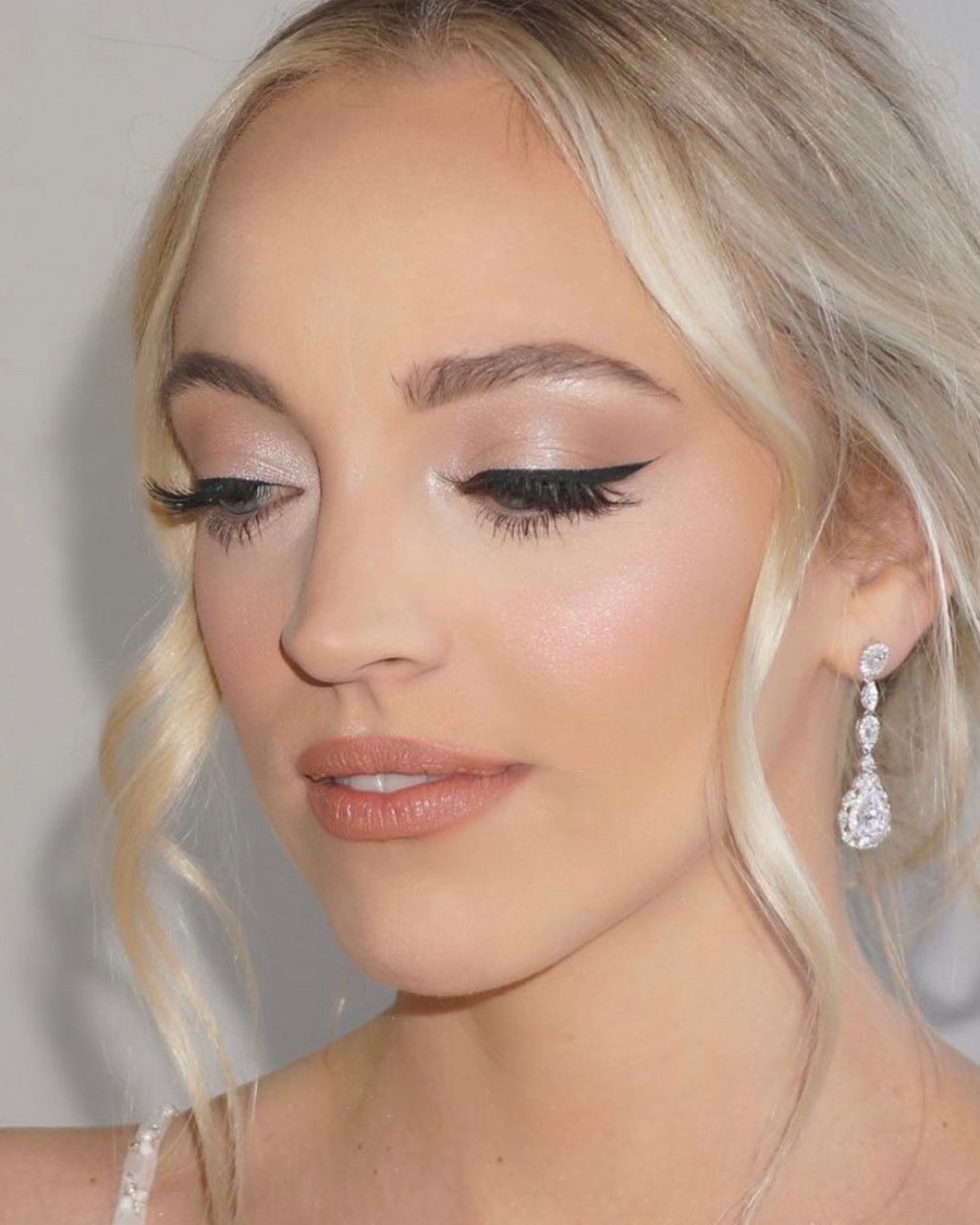 A person with blonde hair and subtle makeup looks down. They have winged eyeliner, light eyeshadow, and wear dangling earrings.