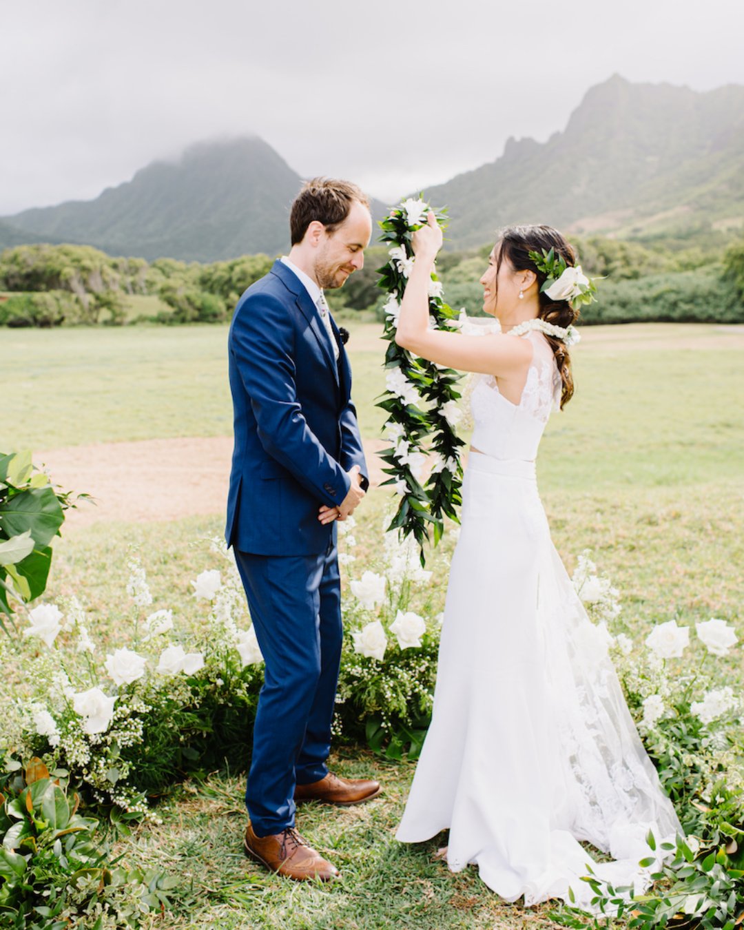 Bride and groom exchanging a floral garland during an outdoor wedding ceremony, surrounded by greenery and mountains.