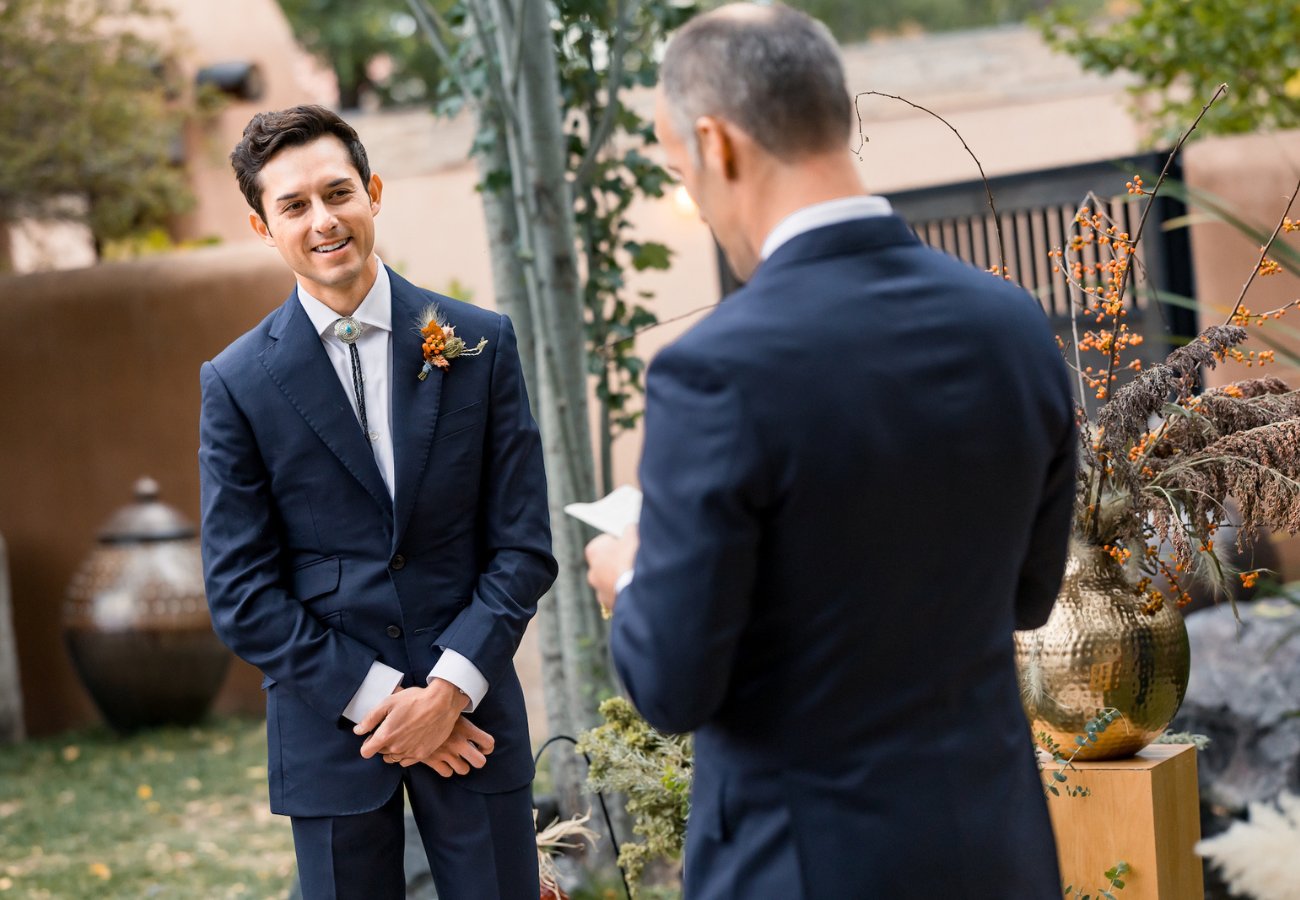 A man in a suit smiles and listens as another man reads from a piece of paper during an outdoor event, possibly a wedding ceremony.