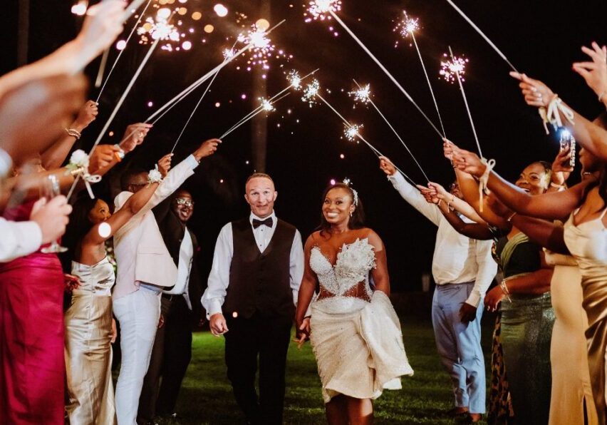 A bride and groom walk hand in hand under a tunnel of sparklers held by guests at an outdoor nighttime celebration.