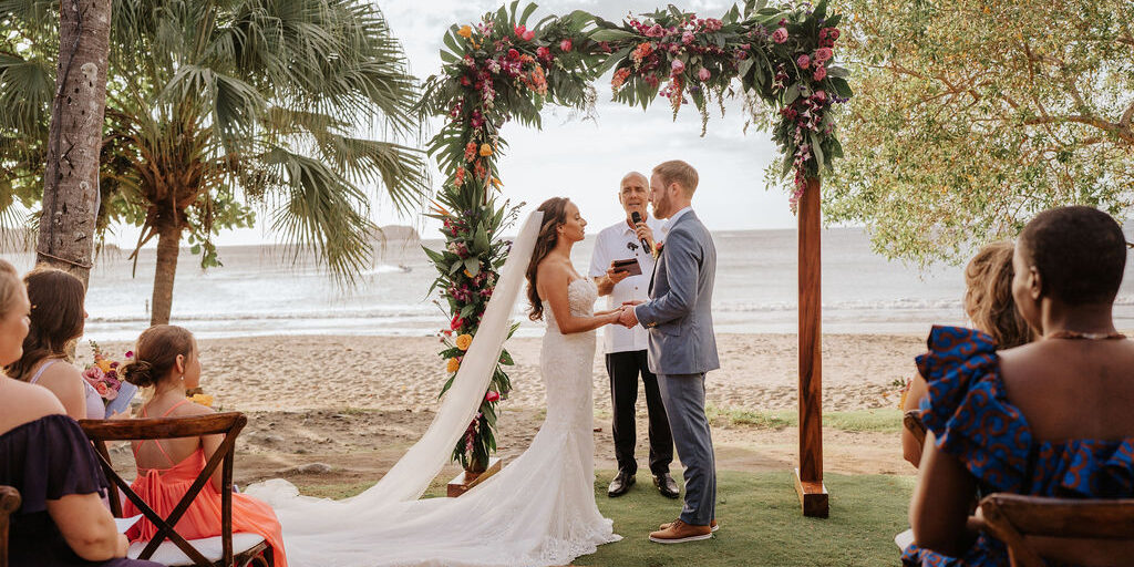 A bride and groom exchange vows under a floral arch at a beachside wedding ceremony, with guests seated nearby and the ocean in the background.