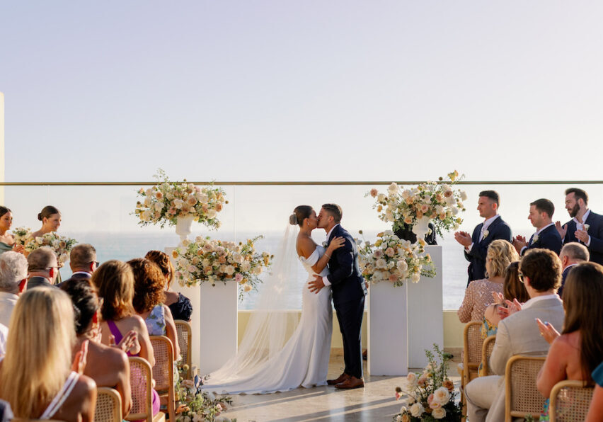 A bride and groom kiss at an outdoor wedding ceremony, surrounded by floral arrangements and guests seated on either side, with a clear sky in the background.