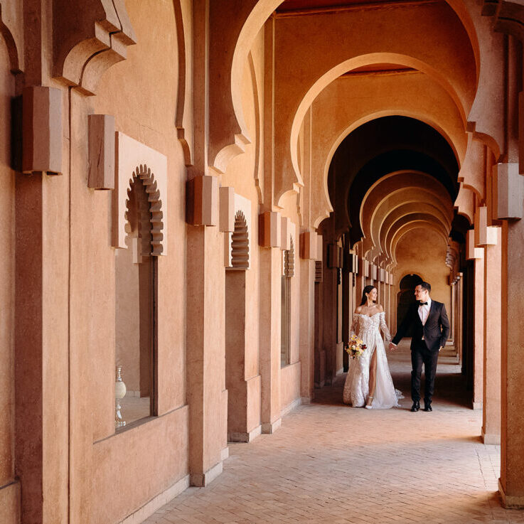 A couple dressed in formal wedding attire walks hand in hand through a corridor with arches and warm-toned walls.