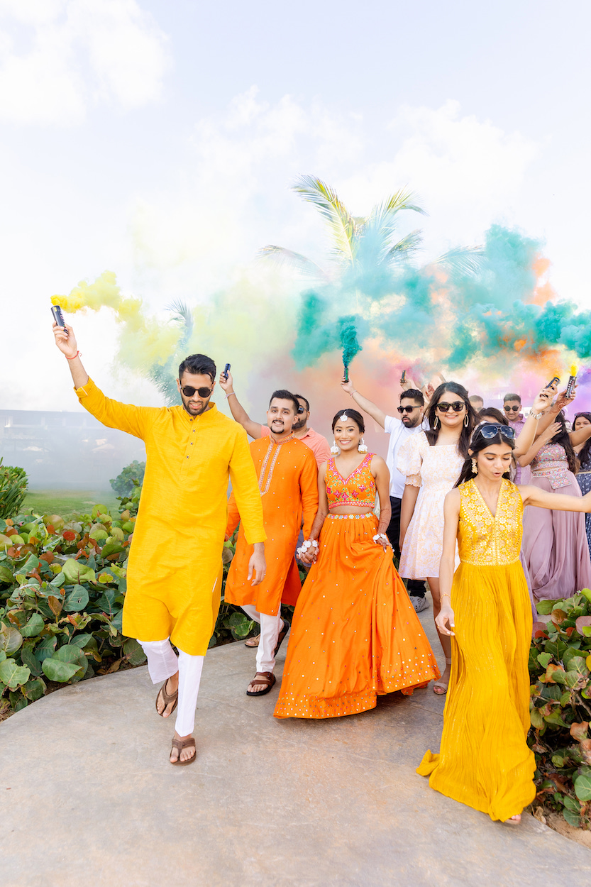 A group of people in bright traditional attire walk outdoors, with colorful smoke in the background.