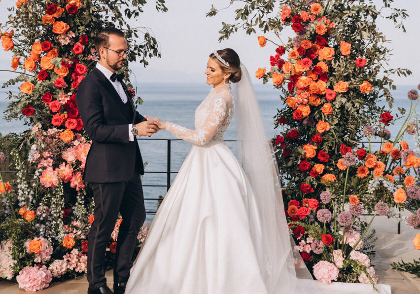 A bride and groom stand facing each other at an outdoor wedding ceremony in front of colorful floral arrangements, with the ocean in the background.