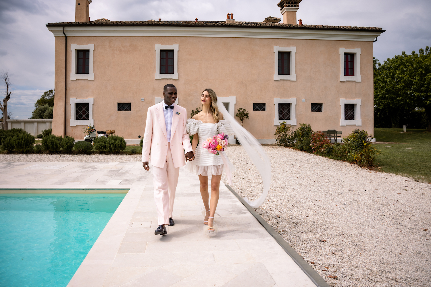 A couple in formal wedding attire walks hand in hand by a pool, with a bouquet and a villa in the background.