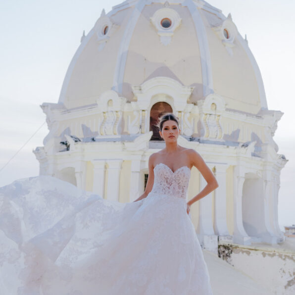 A woman in a strapless white wedding dress stands in front of a large white domed building, holding part of her dress out to the side.