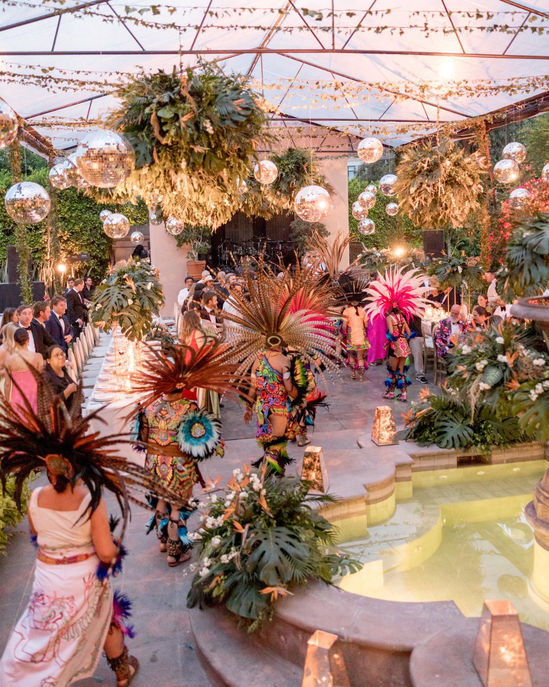 People in traditional Aztec attire perform a dance around a decorated fountain at an outdoor event, surrounded by plants and hanging disco balls.