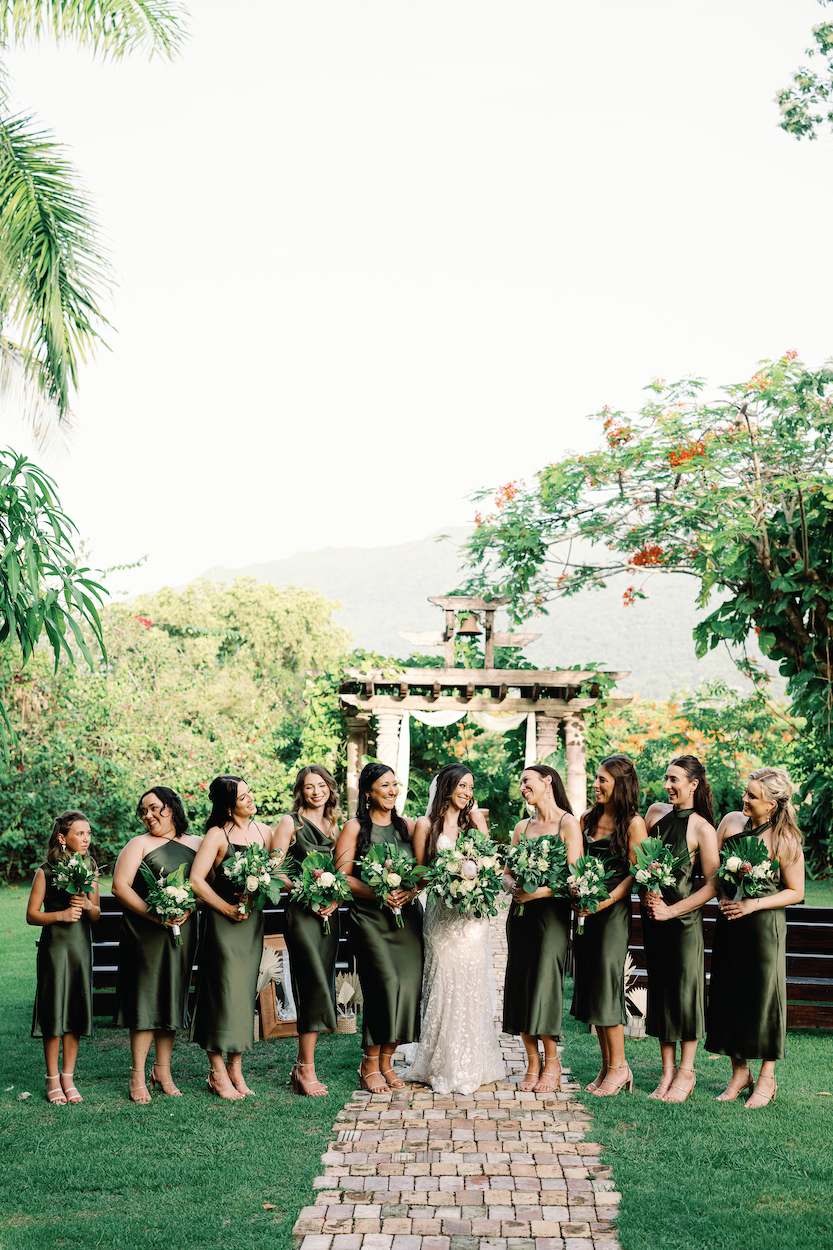 A bride stands outdoors with ten bridesmaids in green dresses, all holding bouquets, on a stone path with lush greenery and a decorated gazebo in the background.