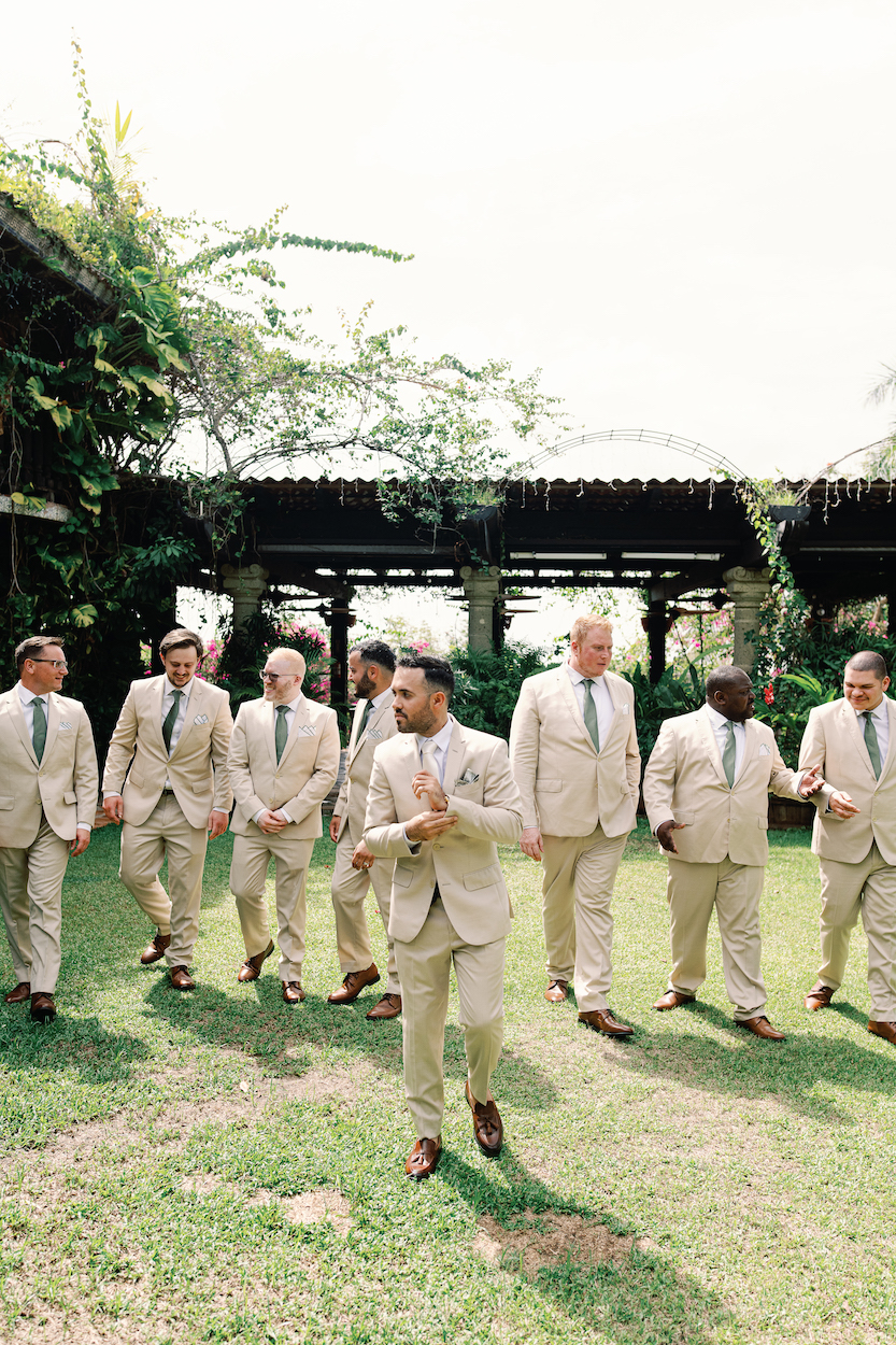 A group of nine men in matching light-colored suits and ties stand and walk on grass in an outdoor garden setting.