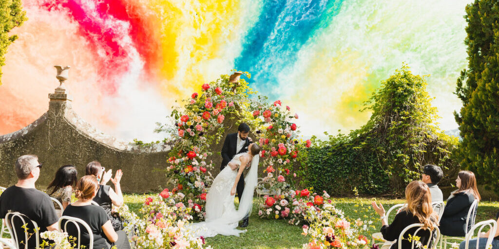 A bride and groom kiss under a floral arch at an outdoor wedding ceremony, with guests seated among flowers and a vibrant, colorful smoke fireworks in the background.