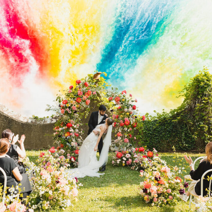 A bride and groom kiss under a floral arch at an outdoor wedding ceremony, with guests seated among flowers and a vibrant, colorful smoke fireworks in the background.