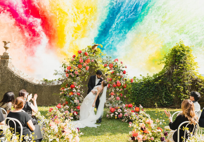A bride and groom kiss under a floral arch at an outdoor wedding ceremony, with guests seated among flowers and a vibrant, colorful smoke fireworks in the background.