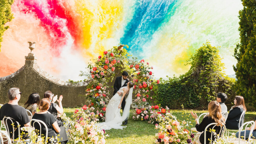A bride and groom kiss under a floral arch at an outdoor wedding ceremony, with guests seated among flowers and a vibrant, colorful smoke fireworks in the background.