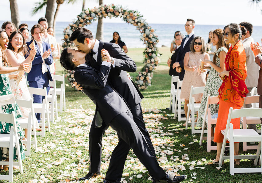 groom dips and kisses his groom with wedding guests clapping to the side and circle floral arch and ocean in background