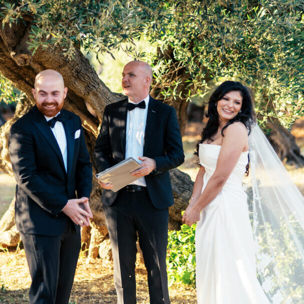 A bride and groom in formal attire stand with an officiant outdoors under a large tree during a wedding ceremony.