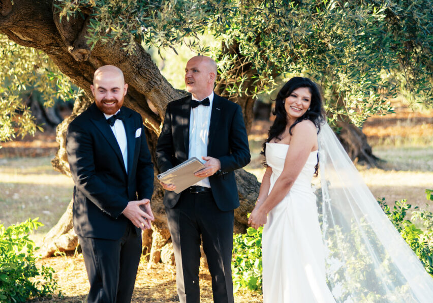 A bride and groom in formal attire stand with an officiant outdoors under a large tree during a wedding ceremony.