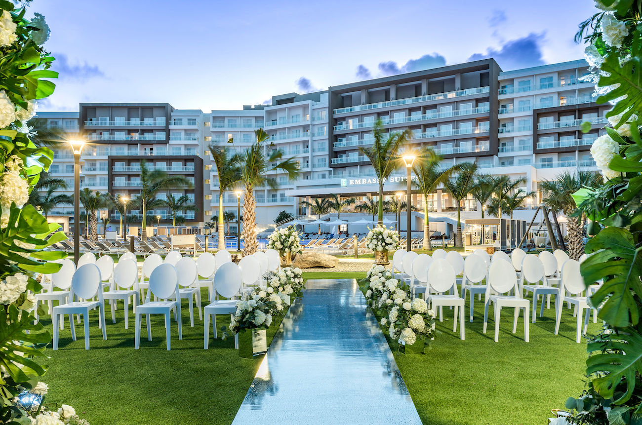 Outdoor wedding ceremony setup with white chairs, floral arrangements, and a blue aisle in front of Embassy Suites by Hilton Aruba Resort with palm trees and a pool in the background.