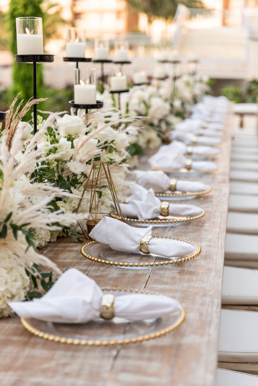 A long wooden table is set with white napkins on gold-rimmed plates, elegant floral arrangements, and candle holders, ready for a formal event.