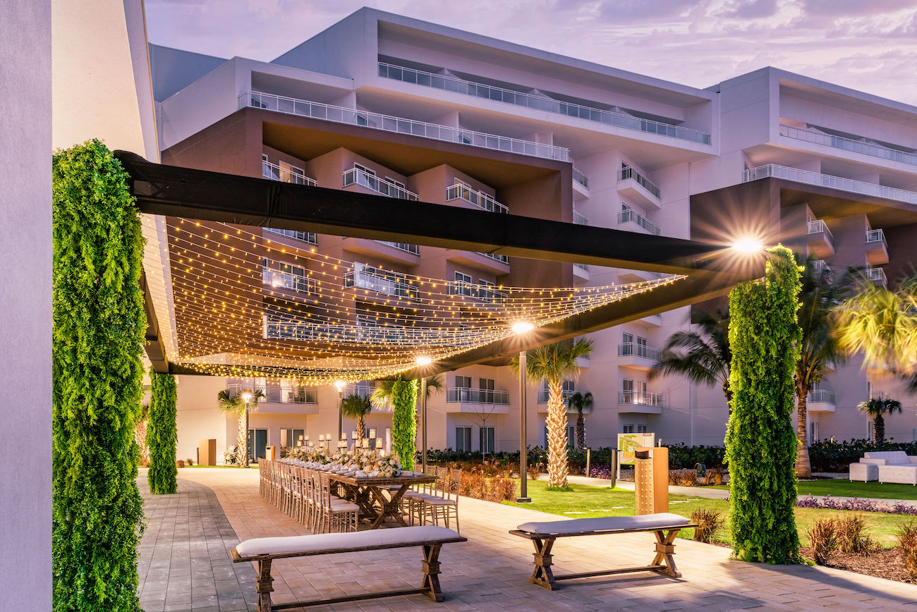 Outdoor dining area with long table and benches under a pergola decorated with string lights, next to a modern multi-story building.