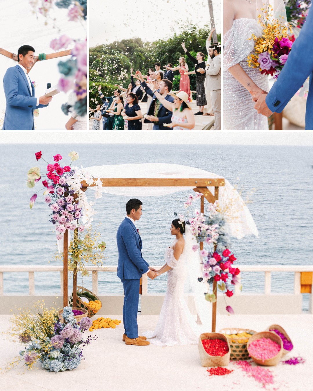 A couple stands under a floral wedding arch by the sea, holding hands, with baskets of flower petals on the ground and guests celebrating in the background.