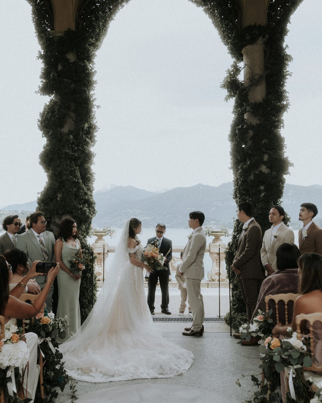 A couple stands under an arch covered in greenery, exchanging vows during an outdoor wedding ceremony. Guests are seated on either side, with mountains visible in the background.