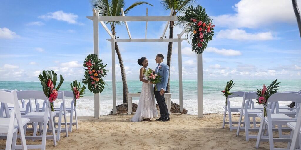 Bride and groom stand under a decorated white pergola on a sandy beach with palm trees, ocean, and flower arrangements in the background.