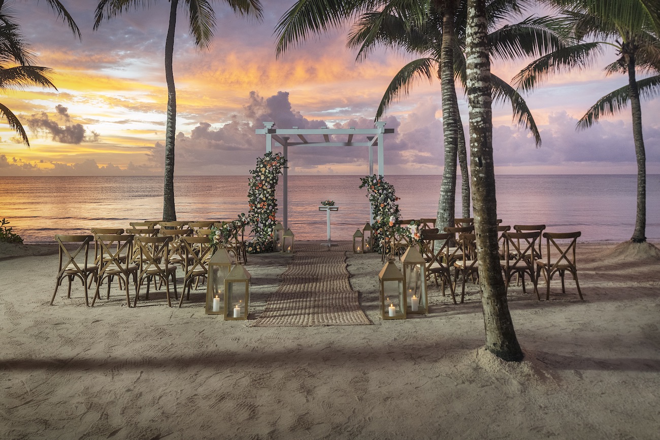 A beach wedding setup with chairs and an arch decorated with flowers, set on sand under palm trees at sunset by the ocean.
