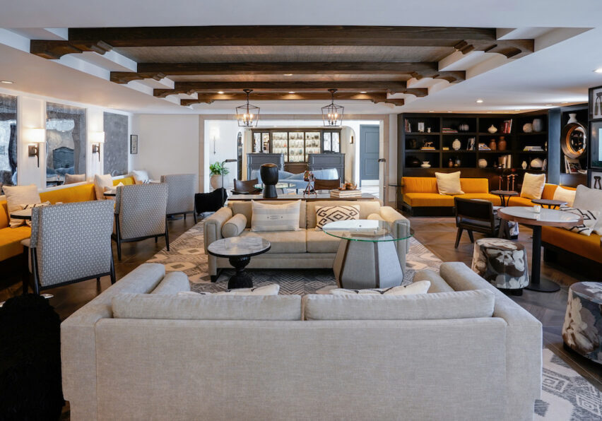 Spacious living room with beige sofas, armchairs, a glass coffee table, wooden ceiling beams, bookshelves, and a yellow couch.