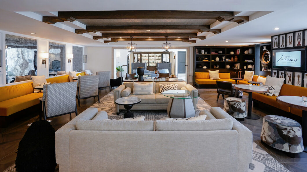 Spacious living room with beige sofas, armchairs, a glass coffee table, wooden ceiling beams, bookshelves, and a yellow couch.