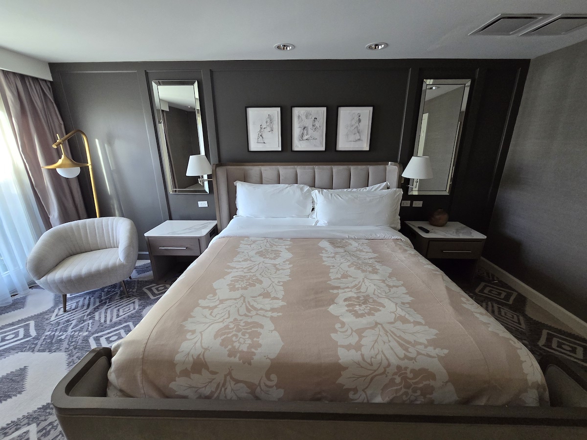A neatly made bed with a beige floral blanket, flanked by two nightstands and lamps, sits against a dark accent wall with three framed pictures; a chair and floor lamp are on the left.