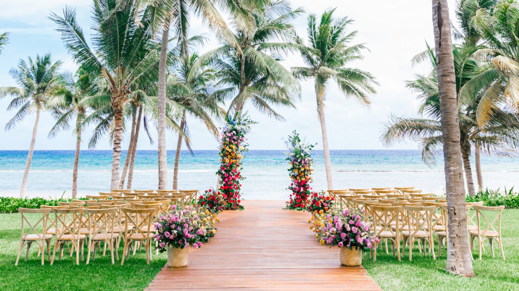 A wooden aisle lined with floral arrangements and chairs leads to two colorful flower pillars on a beach, with palm trees and the ocean in the background.