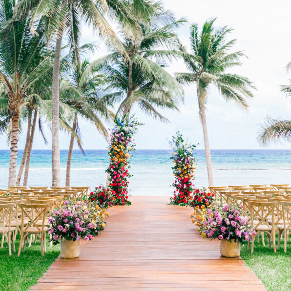 A wooden aisle lined with floral arrangements and chairs leads to two colorful flower pillars on a beach, with palm trees and the ocean in the background.