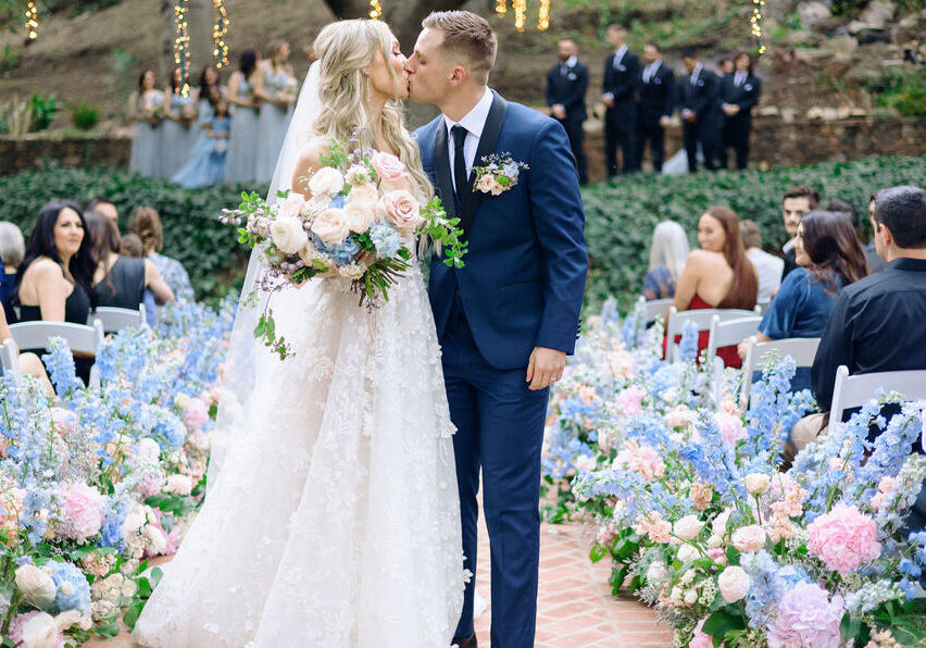 Bride and groom share a kiss at an outdoor wedding ceremony, surrounded by guests and pastel floral arrangements.