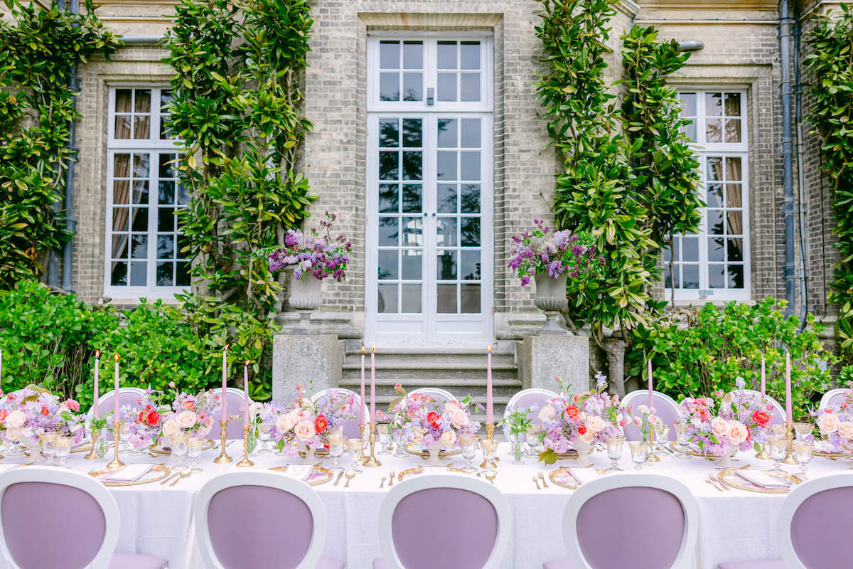 Elegant outdoor dining setup with a long table adorned with pink and purple floral arrangements, candles, and white chairs, set against a brick building with large windows and greenery.