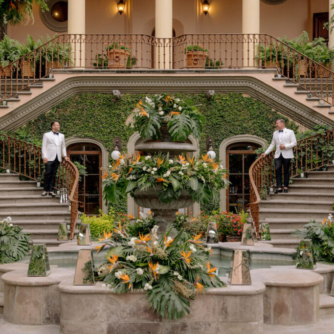 Two men in white jackets stand on opposite sides of a grand staircase behind a large, flower-adorned fountain in front of a building covered with ivy.