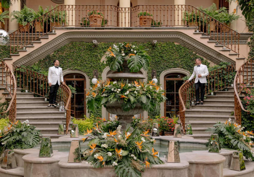 Two men in white jackets stand on opposite sides of a grand staircase behind a large, flower-adorned fountain in front of a building covered with ivy.