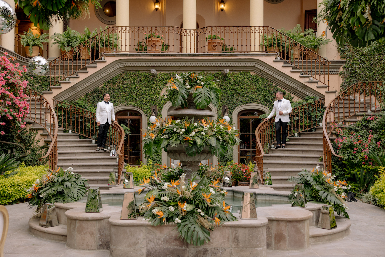 Two men in white jackets stand on opposite sides of a grand staircase behind a large, flower-adorned fountain in front of a building covered with ivy.