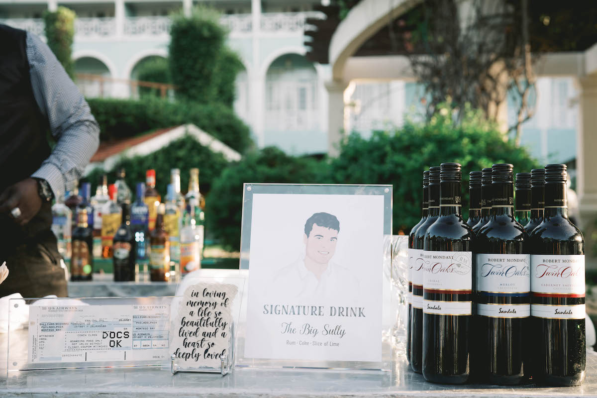 Outdoor bar setup with bottles of liquor, a display sign reading "Signature Drink: The Awk Sorky," bottles of red wine, and a jar with handwritten notes on a table.