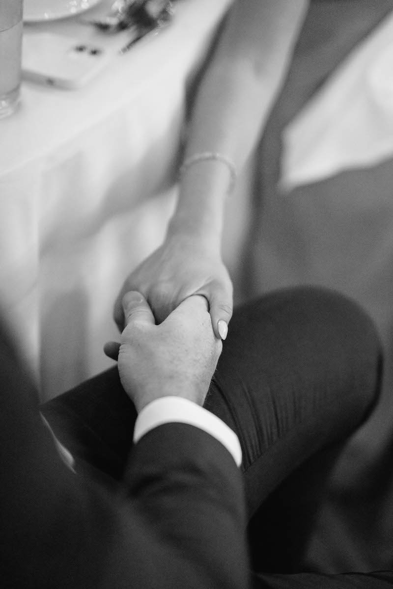 A black and white photo showing two people holding hands across a table; one person is wearing a suit, and only their hands and forearms are visible.