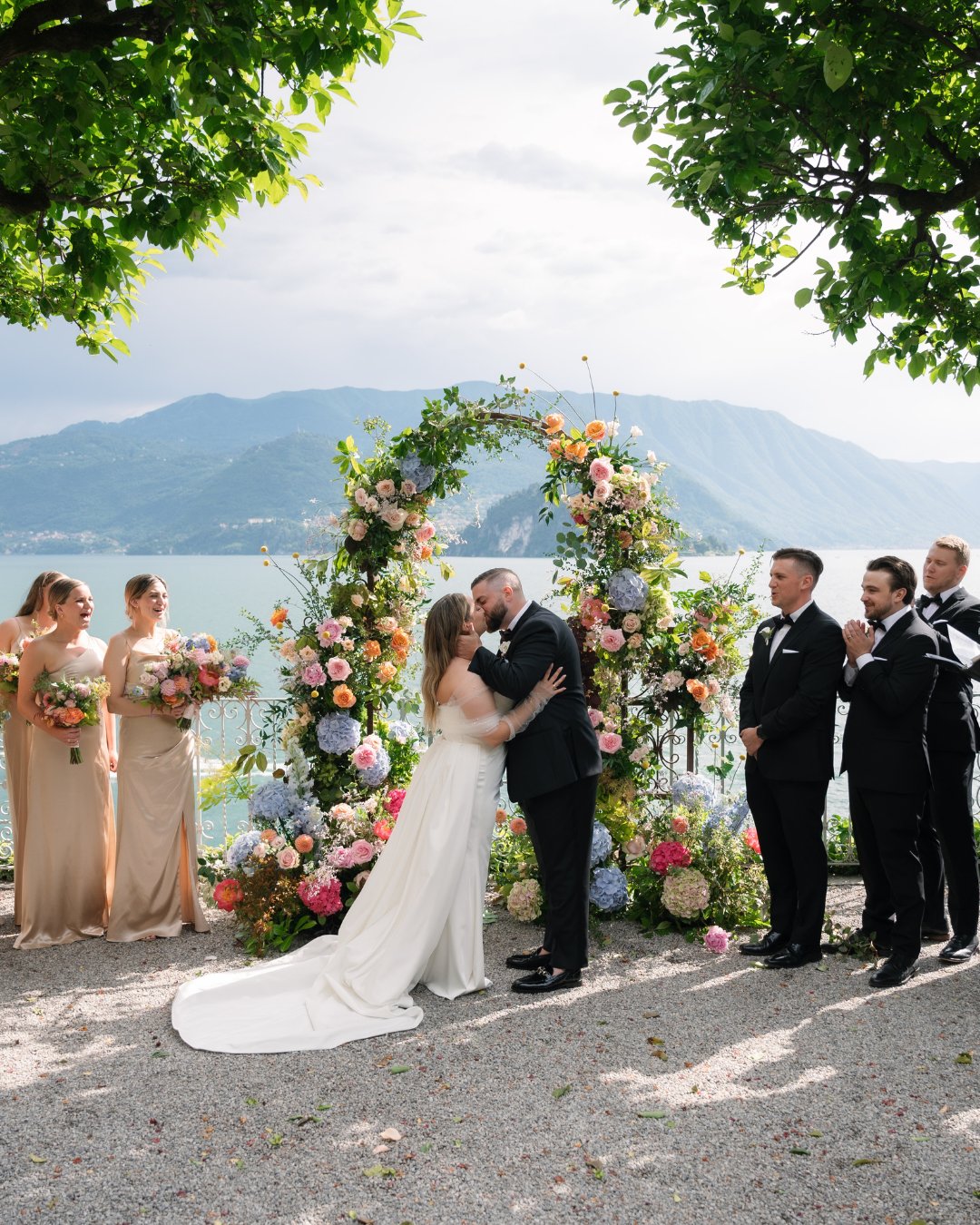 A bride and groom kiss at an outdoor wedding ceremony by a lake, with bridesmaids and groomsmen standing nearby. An arch decorated with flowers frames the couple, and mountains are visible in the background.
