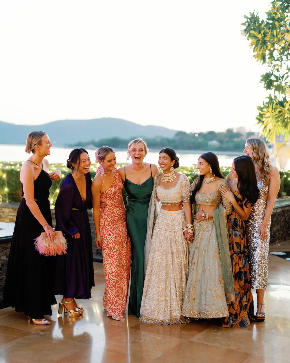 A group of eight women dressed in formal and traditional attire stand together outdoors, smiling and laughing, with a lake and hills in the background.