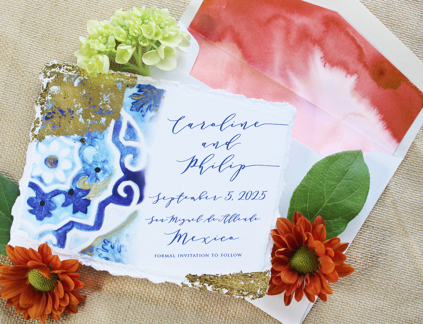Wedding invitation with blue and gold accents. Text reads 'Caroline and Philip, September 5, 2025, Mexico.' Surrounded by flowers and an envelope with a watercolor design.