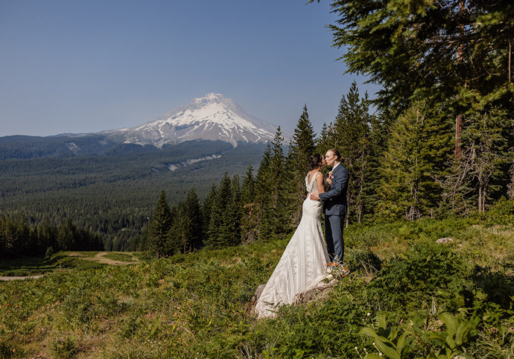 A bride and groom stand on grass surrounded by trees with a snow-capped Mt. Hood in the background under a clear sky.