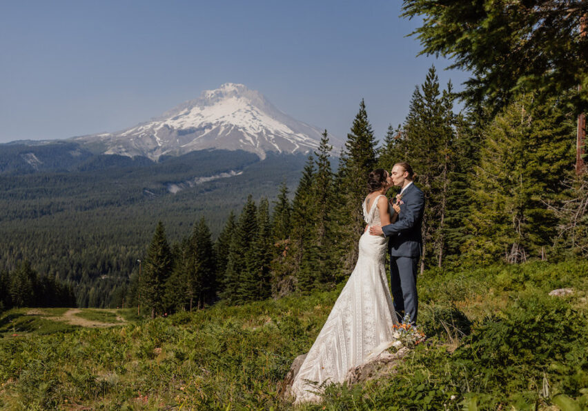 A bride and groom stand on grass surrounded by trees with a snow-capped Mt. Hood in the background under a clear sky.
