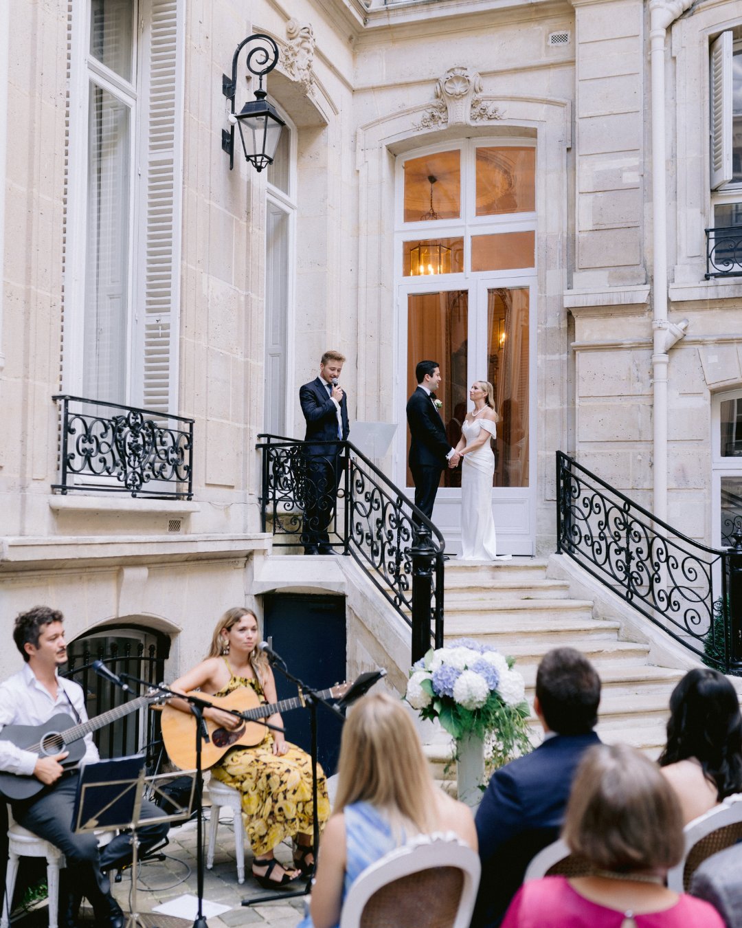 A couple stands on outdoor steps with a man officiating their wedding. Musicians and guests are seated below. The building has ornate iron railings and stone walls.