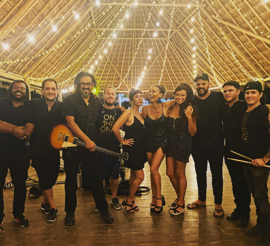A group of musicians dressed in all black smiling under a thatched roof with string lights